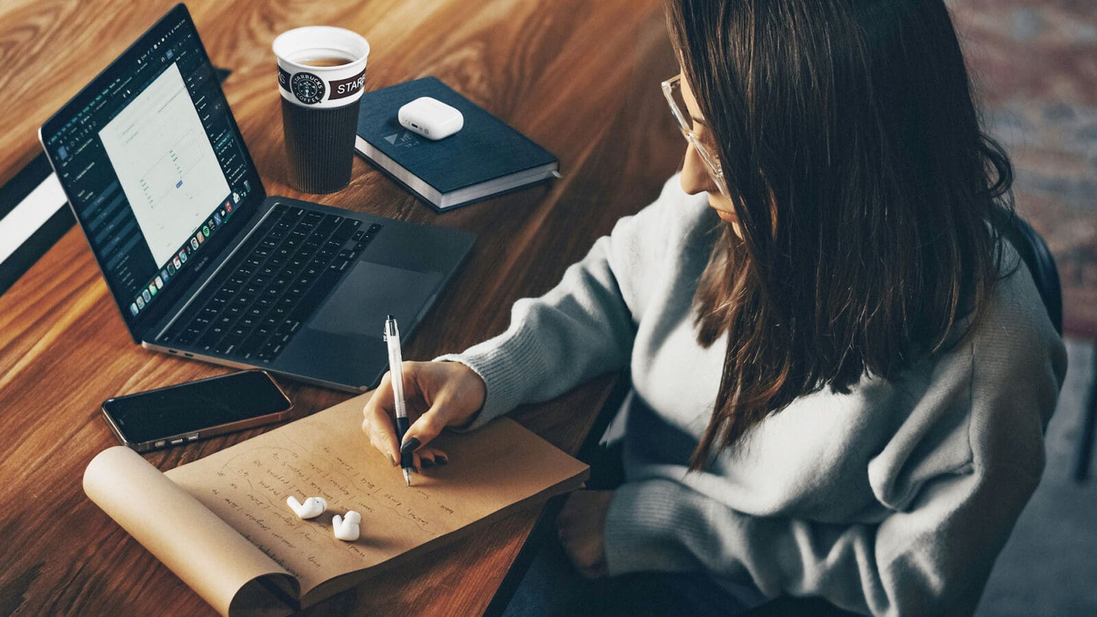 Woman write on notepad next to laptop with airpods and a cup of coffee.