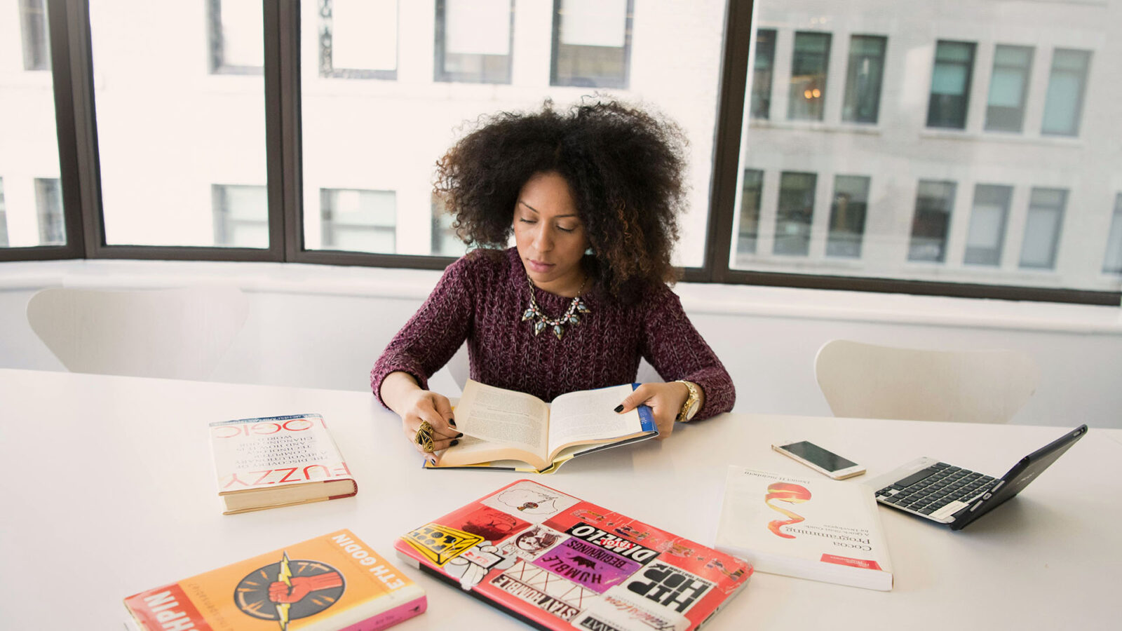Black woman sitting at large table along reading a book surrounded but digital devices and other books.