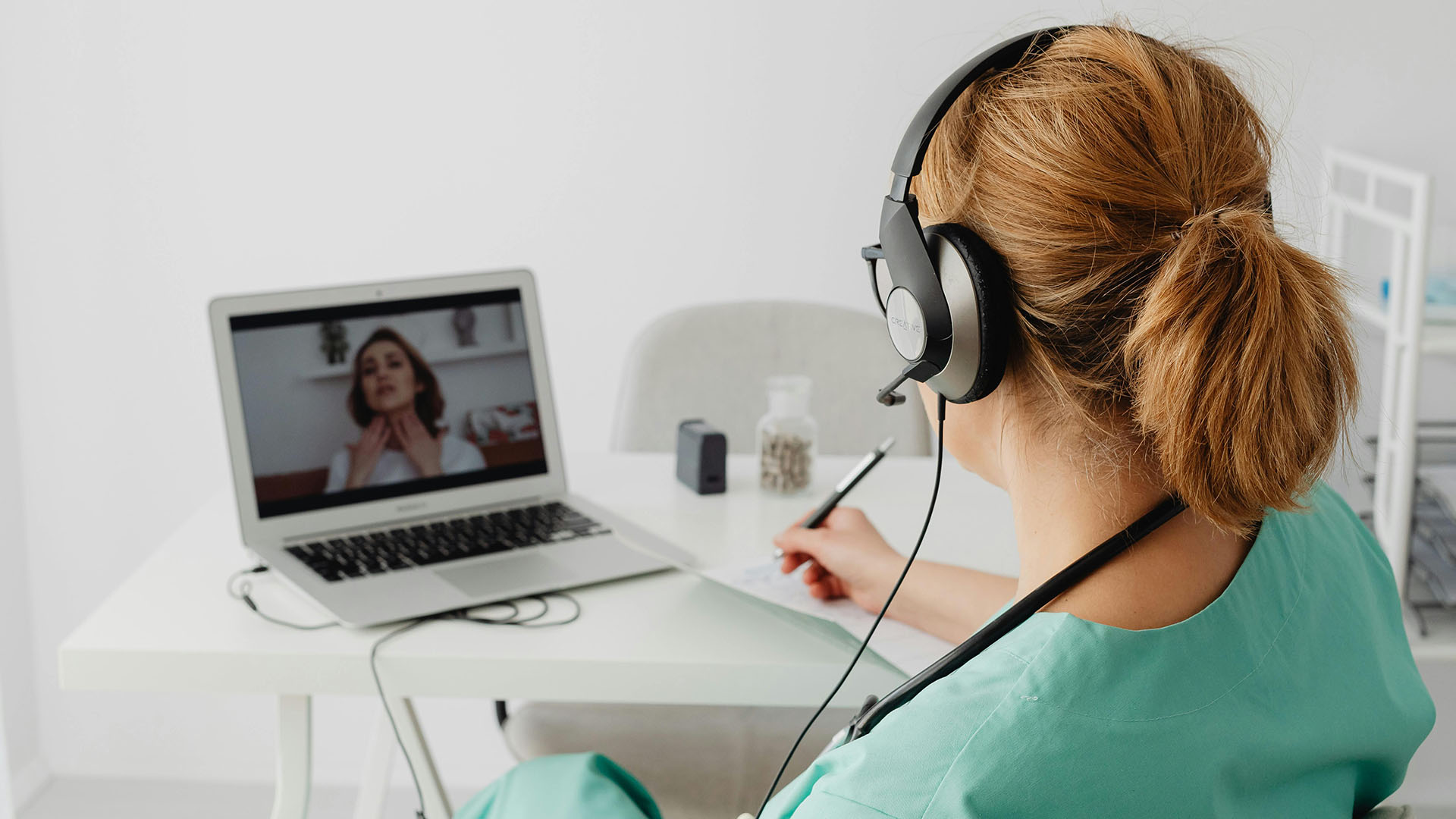 Medical personnel with headphones on facing laptop computer screen with patient pointing to her neck.