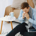 Man sitting in floor leaning against couch sneezing into tissue with tablet in his lap.