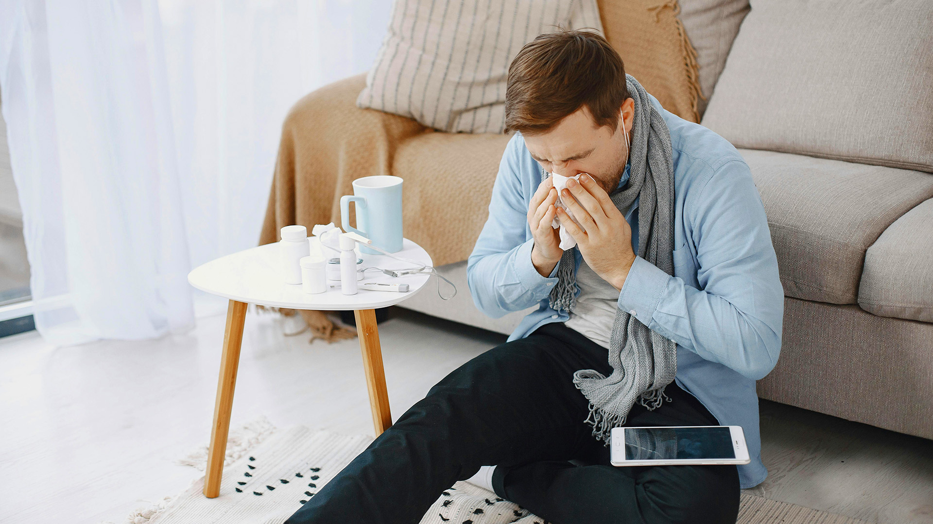 Man sitting in floor leaning against couch sneezing into tissue with tablet in his lap.