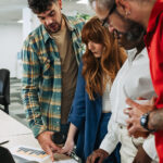 Diverse team working together, they are standing and looking at papers and a computer on a table.