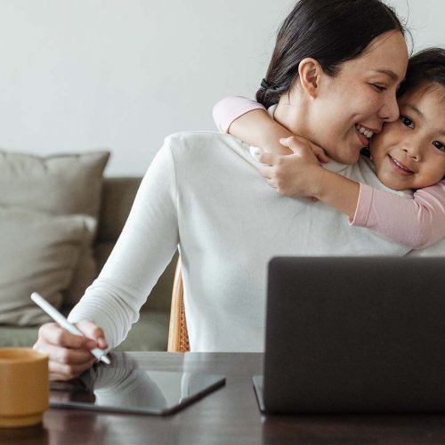 Mother and daughter hugging at table while mother is at computer..